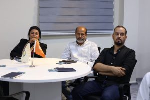 Three adults sit around a round white table in a meeting room, with notebooks and two small flags visible in the foreground.