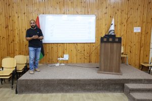 C795A4DD-6BEC-496F-AC9B-741F064FA12A - جامعة سبها | Sebha University Man standing at the front of a lecture room with a whiteboard, podium, and flags nearby, presenting to an audience of empty chairs.