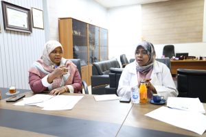 C97BC2AC-A2D5-4A8E-95BD-822E01DD1CB6 - جامعة سبها | Sebha University Two women wearing hijabs sit at a conference table in a modern office, talking with papers and drinks on the table.