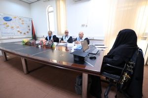 Panel of four men in academic gowns seated at a long conference table; a woman in a black abaya faces them with a laptop nearby.