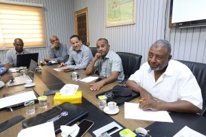 CC83323A-0292-4220-860B-65C29F434F7F - جامعة سبها | Sebha University Five men sit around a conference table in a meeting room with laptops, papers, and coffee cups, looking toward the camera.