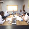 Six professionals sit around a wooden conference table reviewing documents during a meeting in a bright office.