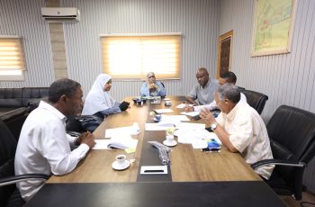 Six professionals sit around a wooden conference table reviewing documents during a meeting in a bright office.