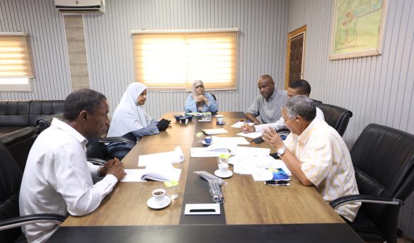 Six professionals sit around a wooden conference table reviewing documents during a meeting in a bright office.