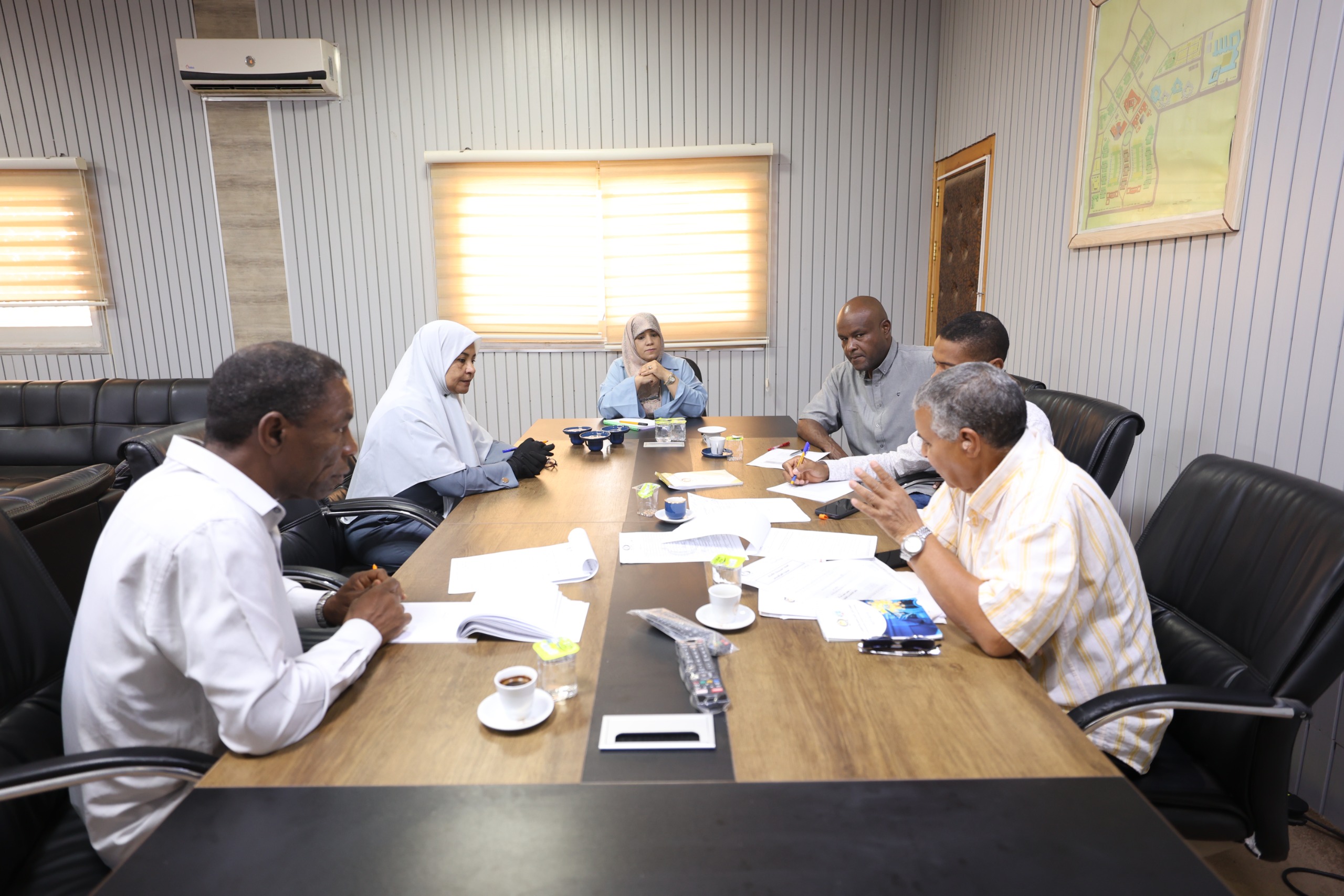 Six professionals sit around a wooden conference table reviewing documents during a meeting in a bright office.