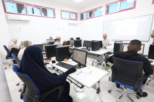 CEA5D72D-EBCF-44B9-8120-50B037D092EB - جامعة سبها | Sebha University A computer lab classroom with students at desks using desktops as an instructor teaches at the front with a projector screen showing a presentation.