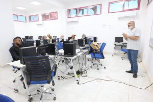 D076BE77-06EB-47EF-8272-A2B18FFB87E6 - جامعة سبها | Sebha University Instructor with gray beard teaching a computer lab; students sit at desks with desktops and cables in a bright room.