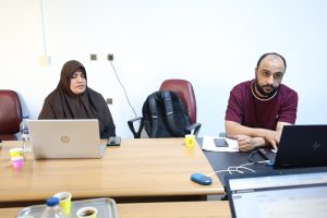 D0ADCEF6-E560-4FEA-97F1-DF1F890C3BB5 - جامعة سبها | Sebha University Two colleagues sit at a wooden table with laptops; woman in a hijab on the left, man with beard on the right.