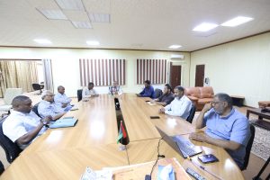 D12E0C46-B329-4255-BB3F-E91EF5647FF9 - جامعة سبها | Sebha University Men in business attire seated around a long conference table with laptops, flags, and documents in a meeting room.”