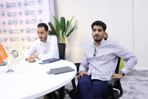 Two men sit at a round white table in a conference room, one in a white shirt and the other in a striped shirt, with notebooks and a backdrop of logos nearby and small flags on the table beside them.