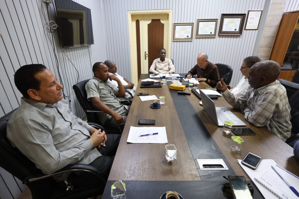 D5437560-F470-4CAD-92D6-E54C748E6895 - جامعة سبها | Sebha University Six men sit around a long conference table in a meeting room, reviewing papers and laptops.