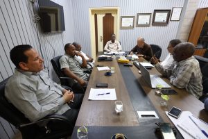 D5437560-F470-4CAD-92D6-E54C748E6895 - جامعة سبها | Sebha University Six men sit around a long conference table in a meeting room, reviewing papers and laptops.
