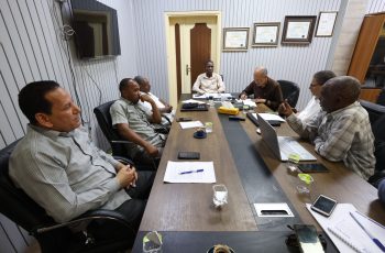 Six men sit around a long conference table in a meeting room, reviewing papers and laptops.