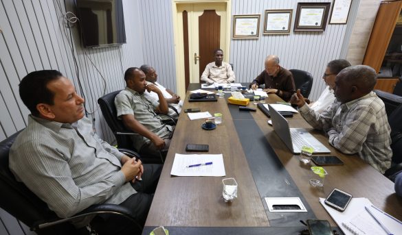 Six men sit around a long conference table in a meeting room, reviewing papers and laptops.