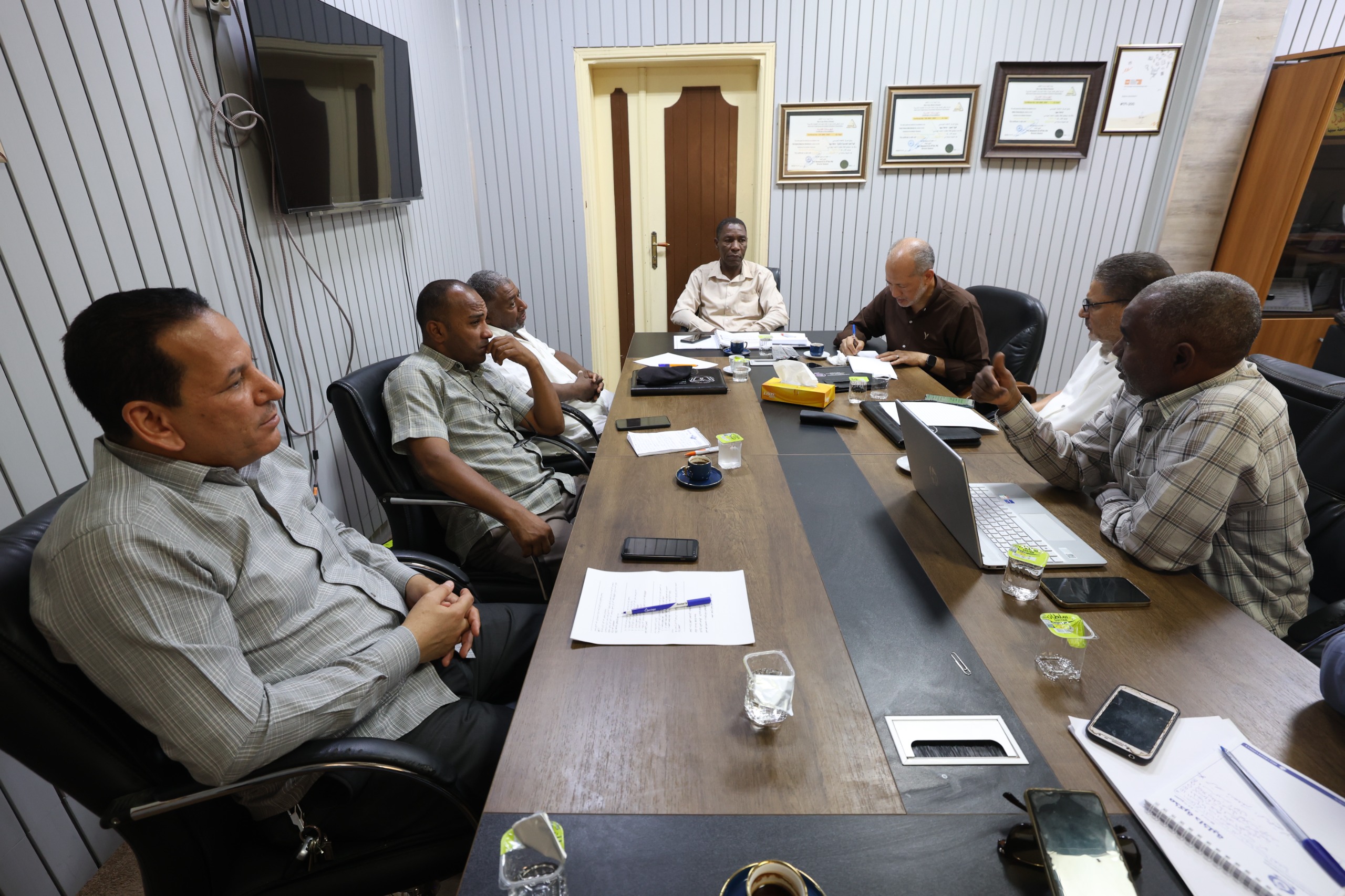 Six men sit around a long conference table in a meeting room, reviewing papers and laptops.
