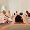 Five men sit around a U-shaped conference table in a bright room, reviewing documents and taking notes.