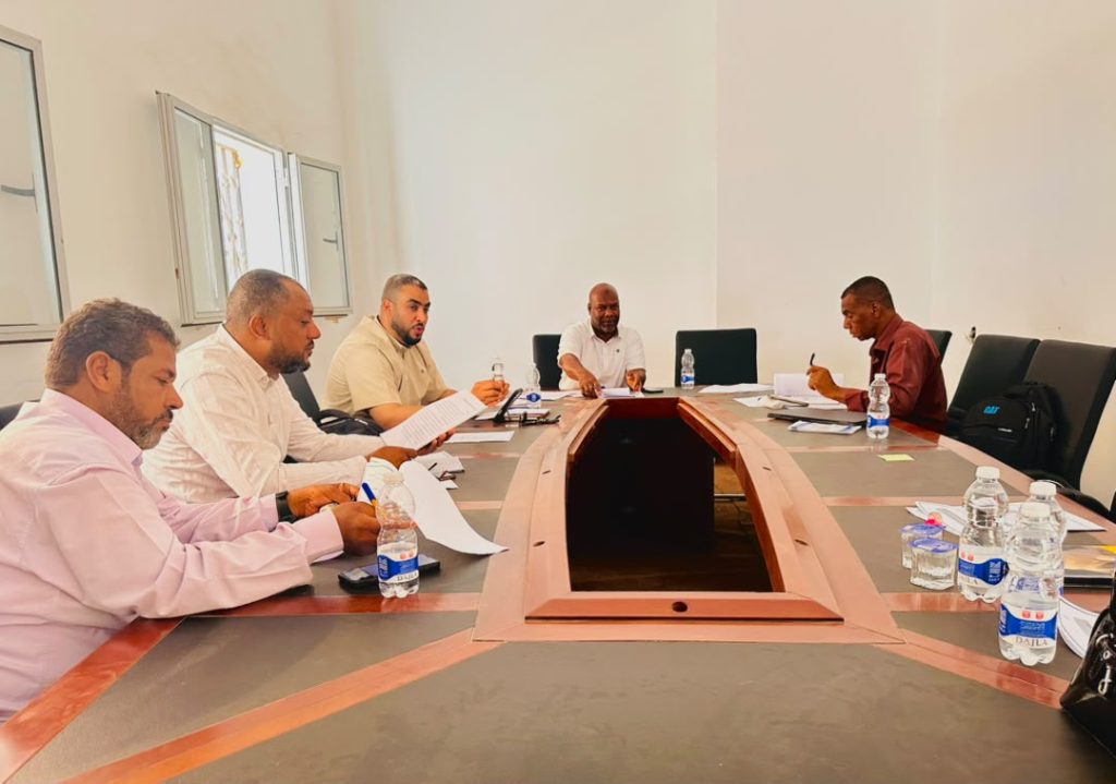 D6585992-6E3F-4B44-9F99-4CD6E1B0CF49 - جامعة سبها | Sebha University Five men sit around a U-shaped conference table in a bright room, reviewing documents and taking notes.