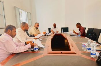 Five men sit around a U-shaped conference table in a bright room, reviewing documents and taking notes.