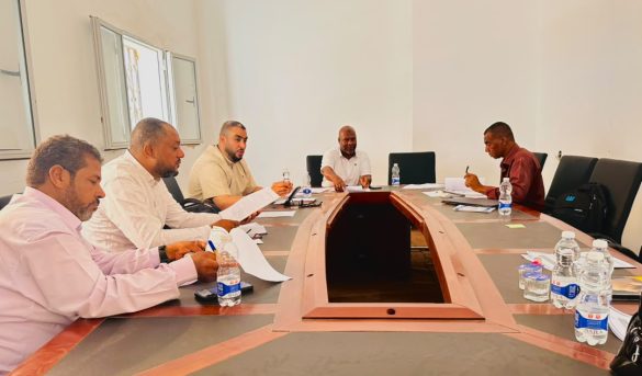 Five men sit around a U-shaped conference table in a bright room, reviewing documents and taking notes.