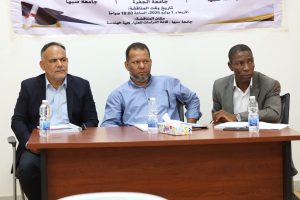 D8F38BE2-E5C6-4375-85B3-FF97FE10B899 - جامعة سبها | Sebha University Three men in business attire sit at a conference table during a panel, with water bottles and a tissue box on the table and an Arabic banner in the background.
