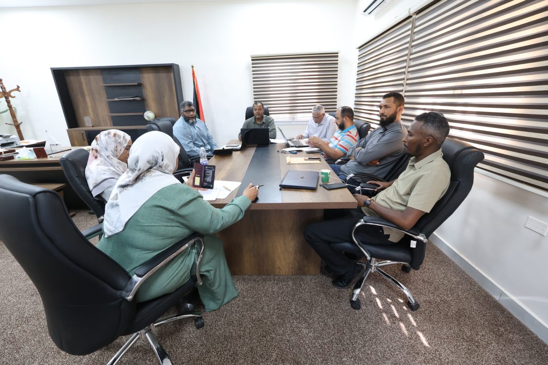 Seven people around a rectangular conference table in a bright office, with laptops, papers, and a water bottle during a meeting.