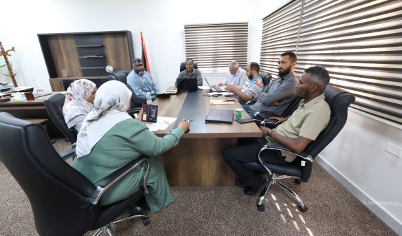 Seven people around a rectangular conference table in a bright office, with laptops, papers, and a water bottle during a meeting.