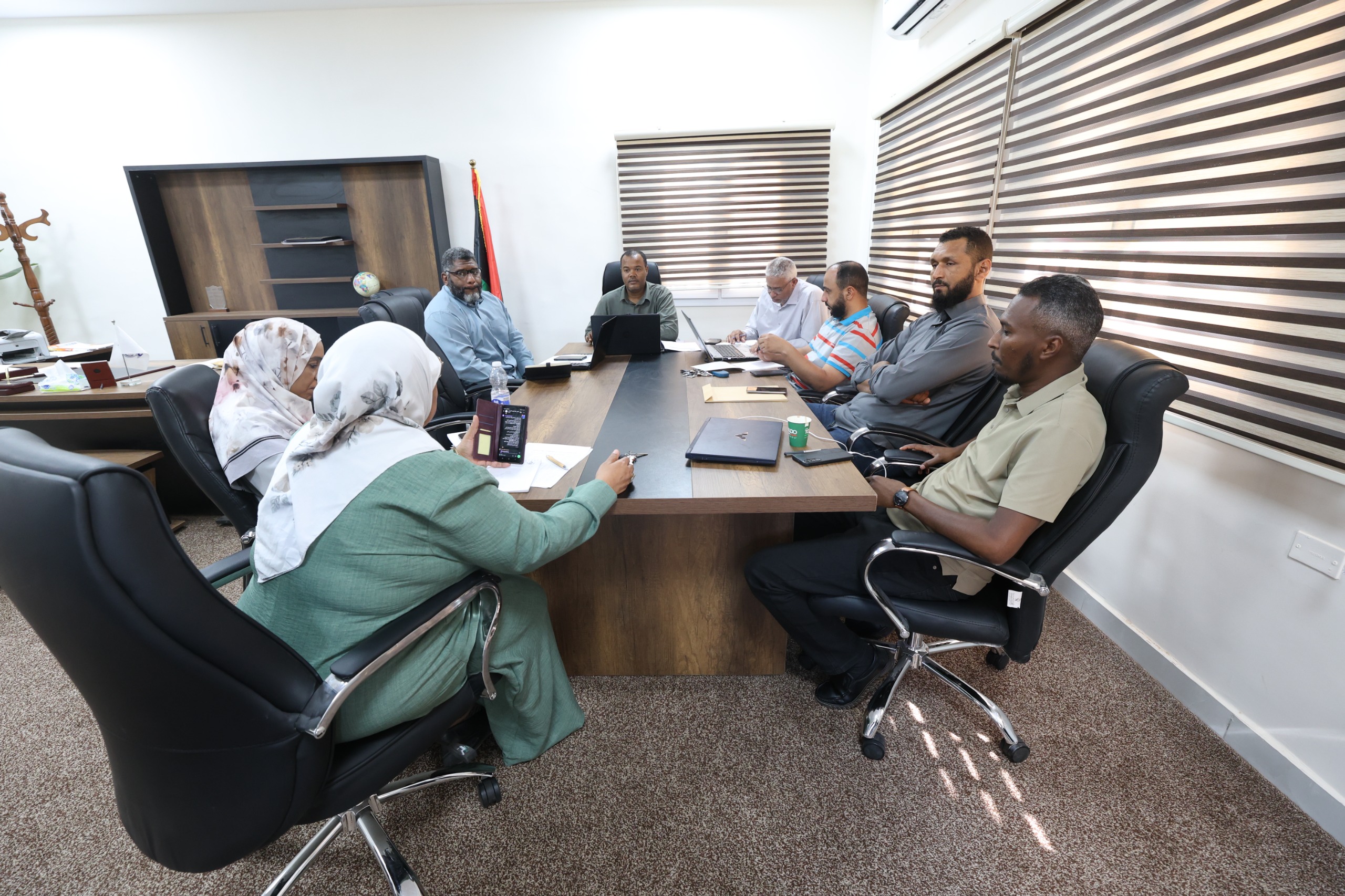 Seven people around a rectangular conference table in a bright office, with laptops, papers, and a water bottle during a meeting.
