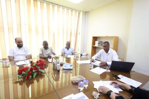 DF54414A-2987-46B8-BB0F-CC4CA023D8B4 - جامعة سبها | Sebha University Four men sit around a glass conference table during a meeting, with papers, drinks, and a Dell laptop visible; a bookshelf sits in the background.