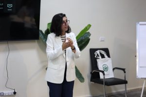 Woman in a white blazer giving a presentation, gesturing with her hands in a conference room.