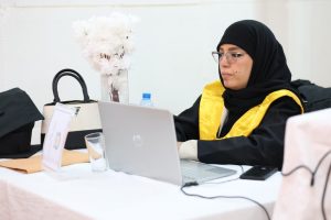 Woman in a black hijab and yellow graduation stole typing on a silver laptop at a table with a water bottle and bag nearby.