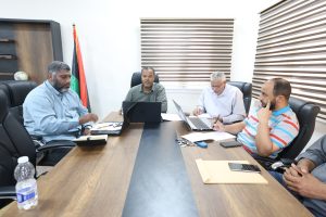 E4248A90-141A-4F0D-B3CC-227E961BAF76 - جامعة سبها | Sebha University Four men sit around a wooden conference table with laptops and papers in a bright office.