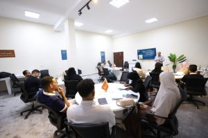 A presenter speaks at a podium in a bright conference room to a round-table group of attendees seated with laptops and notebooks.