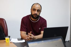 EA402B11-5035-43B5-A009-A371DAE5D3DD - جامعة سبها | Sebha University Man with a beard sits at a conference table looking thoughtful, with a laptop, phone, papers, and a yellow cup nearby.