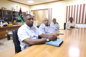 EC4F0174-449B-456F-AB57-683C062D30D6 - جامعة سبها | Sebha University Four men seated at a wooden conference table in a meeting room; Libyan flag visible in the background.