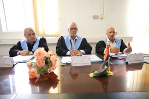 Three men in black robes with light blue sashes sit at a conference table with documents, a flower arrangement, and a small flag on the right.