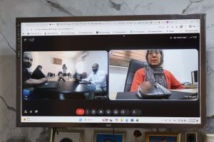 Screenshot of a video conference on a monitor: left pane shows several people around a table with laptops; right pane shows a woman in a hijab at a desk with a mouse.