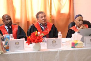 Three judges in red-trimmed gowns sit at a panel table with nameplates, flowers, and water bottles in front of them.