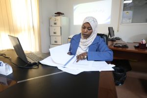 FA61C774-8B71-4783-9EB1-31C01495F832 - جامعة سبها | Sebha University Woman in a blue blazer and hijab sits at a desk, sorting through documents beside a laptop in an office.