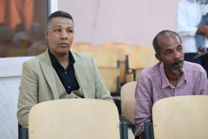 Two men sit in a classroom or conference room, attentively listening; the man on the left wears a beige blazer over a black shirt, the man on the right wears a pink button-down shirt with a gray beard.