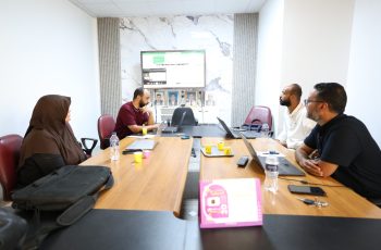 Four people around a wooden conference table with laptops and water bottles, watching a wall-mounted monitor.