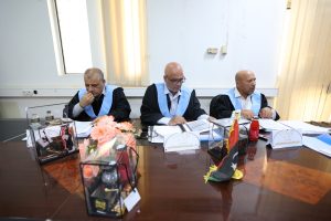 Three judges in black robes with light blue sashes sit at a conference table, reviewing documents, with pink flowers and gift boxes in the foreground.