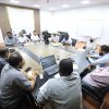 Group of professionals seated around a large conference table in a meeting room, some with laptops and papers in front of them.