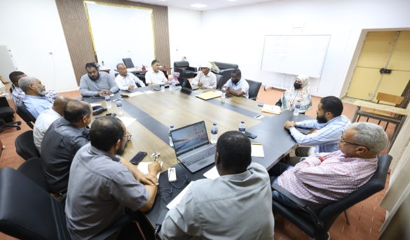 Group of professionals seated around a large conference table in a meeting room, some with laptops and papers in front of them.
