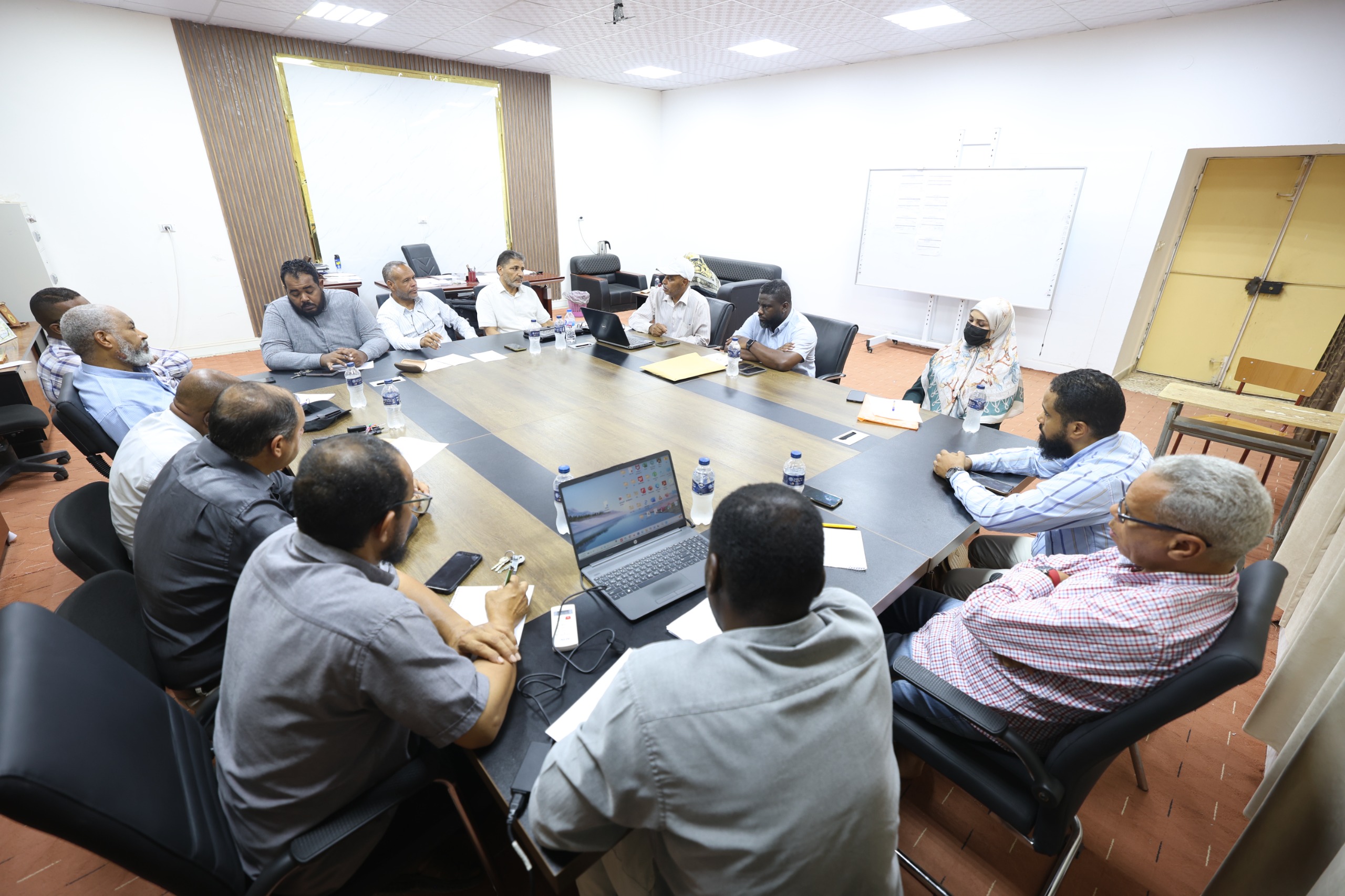 Group of professionals seated around a large conference table in a meeting room, some with laptops and papers in front of them.