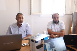 Two men sitting at a meeting table with laptops in a bright office environment.