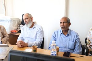 Two middle-aged men seated at a conference table in a bright office; one with a gray beard in a striped shirt and the other in a blue shirt, with cups and a water bottle on the table. How to cite this: the two men are engaged in conversation during a meeting in a well-lit room.