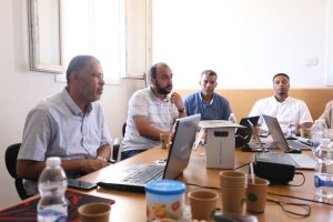 Group of men in a conference room seated around a table with laptops and coffee cups everyonelooking attentive at a discussion.