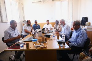 Group of men seated around a conference table in a bright office, working on laptops and sharing snacks during a meeting.