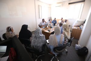 Group of diverse professionals seated around a table in a meeting, with laptops, papers, and drinks as they discuss.]},