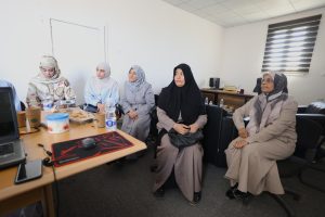 Five women wearing hijabs sit around a conference table in a bright office, with snacks and drinks on the table.